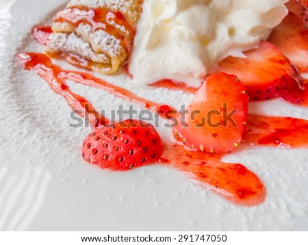 The closeup of slice strawberries put on a white plate with crepes, icing sugar, strawberry sauce and cream.