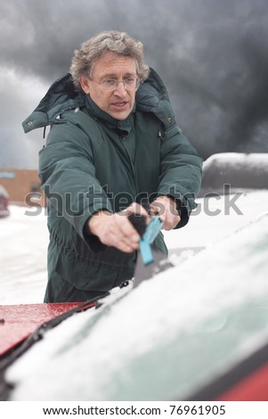 A man is using an ice scraper to scrape off the snow and ice from his car\'s windshield. Vertical shot.