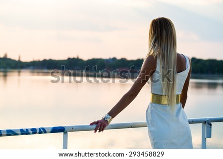 pretty dressed young girl looking to the lake from handrail at sunset