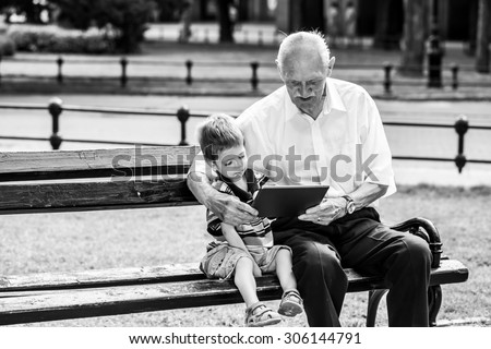 Grandchild teaching to his grandfather to use tablet on a bench. Black and white photography