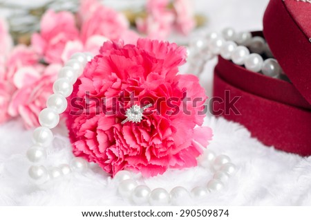 Engagement diamond ring in pink carnation flower with pearl neklace and red jewel box on white fur background