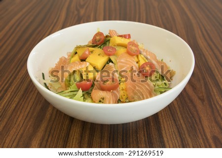 Salad of slices raw salmon and roe on fresh green vegetable with cucumber, cherry tomatoes, avocados and mangoes.  All ingredients are prepared and placed in layers in a large round white bowl.