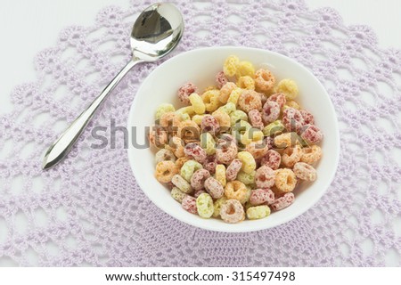 A large white ceramic bowl of colorful breakfast cereal of mixture flavor with a silver spoon placed on a purple lace crochet table cloth.  White background