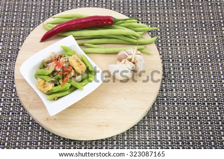 A Chinese cooking dish of saute green string beans with meat, bean curd, onion, garlic and garnished with finely sliced chili. All on a wooden chopping board with brown woven table mat background.