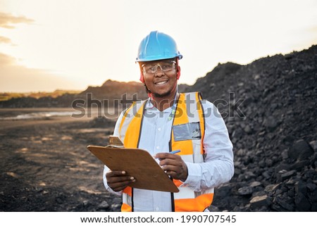 A young Black African coal mine foreman looking into camera smiling wearing reflective bib and hard hat is inspecting samples of coal on his clipboard after a long day of work on site at the coal mine Stock foto © 