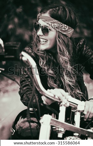 Inked black and white picture of a happy beautiful young biker girl and her motorcycle. Color effects, a soft film noise and a custom white balance is added to this image.