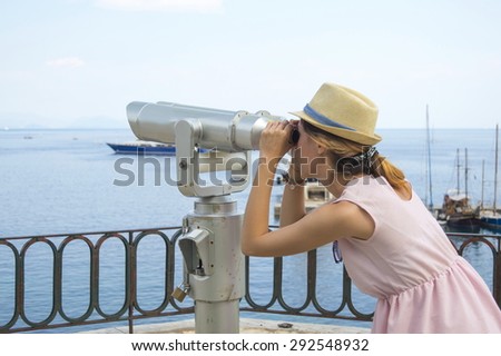 Young girl looking thru public binoculars at the seaside wearing straw hat and pink dress