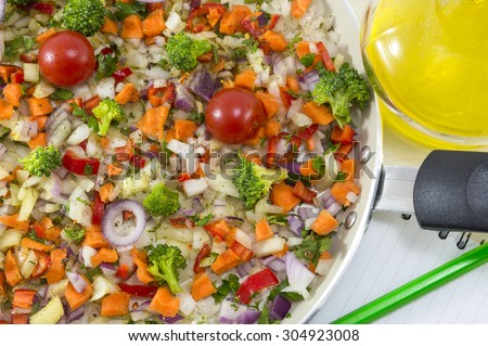 Vegetable salad on a cooking pan next to a open notebook and a pen for writing recipe. Vegetables ready to be cooked with natural  olive oil