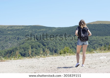 Young, beautiful girl with a backpack on her back, standing on the plateau. Green meadows and majestic mountains in the background. Hiking trip background