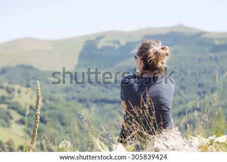 Young woman with backpack sitting on cliff\'s edge and looking to mountains and clear sky. Summer hiking trip adventure
