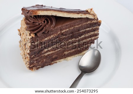 A slice of chocolate cake with chocolate glossy syrup topping and chocolate flower and nuts and a spoon on a white plate on white background