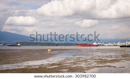 Groups of people wading and playing on the low tide waters of English Bay in Vancouver, with some large ships anchored in the background, on May 5, 2014