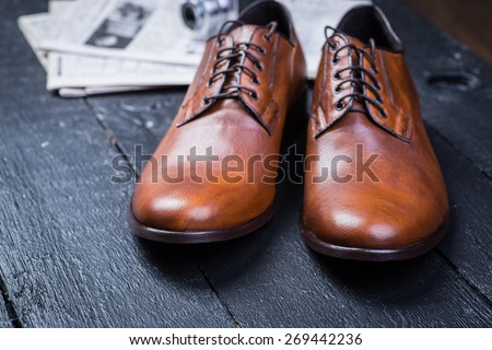 A pair of brown leather shoes with vintage camera and newspaper on a black wooden floor