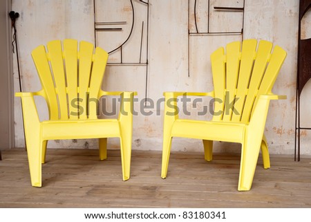 Two yellow plastic chairs on a veranda against an wall with metal art