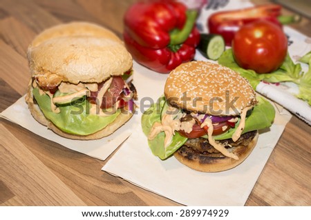 Comparison of two homemade burgers. One hamburger with sesame roll and another with homemade big bun. Background with pepper, tomato and other vegetables on a wooden desk