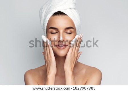 Happy young woman with bath towel on her head takes care of her skin face, applies cleansing foam after shower, smiling and closed eyes, isolated on grey background. Face wash. 