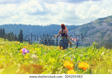 Happy young red hair woman running and dancing on the flowers field into the mountains. Happy travel concept. Mountain trekking. Woman in harmony with nature.