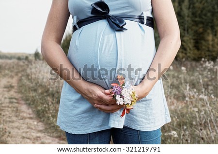 Young pregnant woman in field with a bunch of wild flowers under belly. Selective focus.