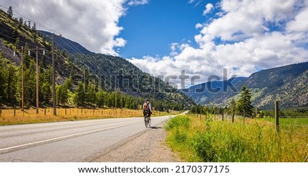Cyclist riding Mountain Bike on highway on sunny summer day in Canada. A lone cyclist on a mountain highway toward distance mountains. Travel photo, summer activity-June 6,2022-Hedley BC, Canada Foto stock © 
