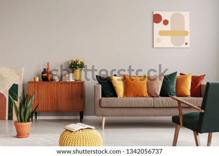 Abstract painting on grey wall of retro living room interior with beige sofa with pillows, vintage dark green armchair and yellow pouf with book