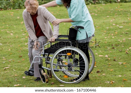 Nurse supporting elder disabled woman to sit on wheelchair
