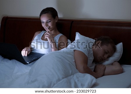 A man sleeping and his woman using a laptop at night in bed