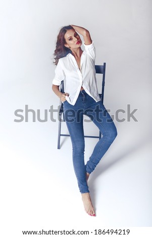 Fashion model sitting on a chair in a blouse and jeans barefoot on a white background