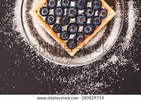 Blueberry tart on a glass saucer on black background and spilling sugar