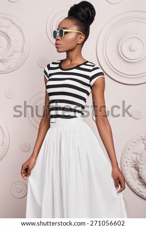 Black girl with clear skin , wearing glasses , a striped T-shirt and long white skirt with a gathered black hair standing on the background of a decorative wall plaster decorated with beige circles