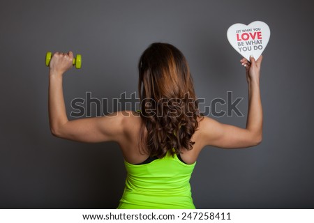 Rear view of beautiful young fitness model (bodybuilder) woman training with dumbbell (weight) and holding a white heart shape with \