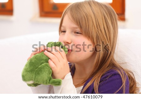 Young girl with blonde hair and purple dress sitting on sofa kissing a toy frog, selective focus on lips
