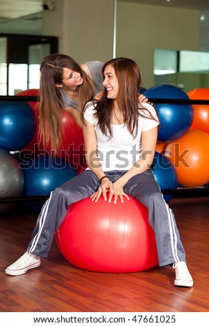Two happy young women in the gym sitting on a red ball smiling to the camera