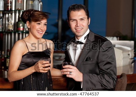 Young couple at a bar, men in black tuxedo holding whiskey in his hand, woman with hairstyle in black dress smiling to the camera