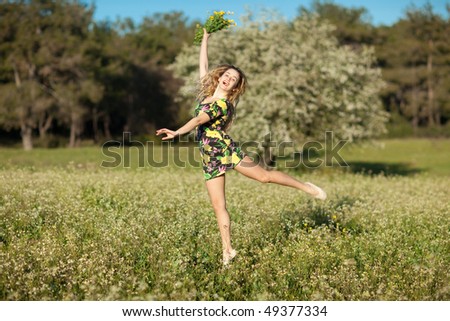 Beautiful young blonde woman jumping in blooming meadow in spring, bunch of yellow flowers in hand; shallow depth of field, trees in background