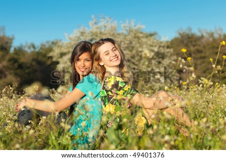 Two beautiful young women sitting in blooming meadow in spring, smiling, blue sky and trees in background; shallow depth of field