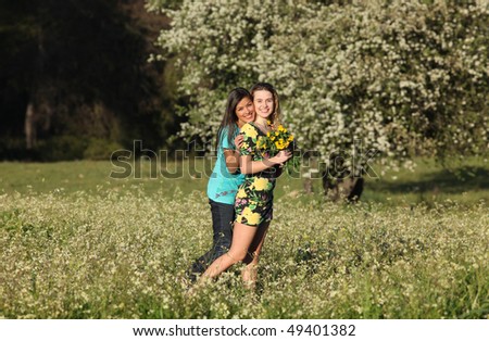 Two beautiful young women standing in blooming meadow in spring, smiling, trees in background; shallow depth of field