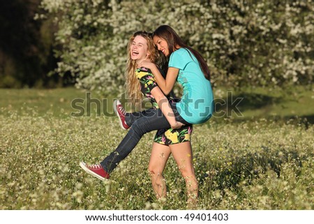 Two beautiful young women standing in blooming meadow in spring, giving piggyback, smiling, tree in background; shallow depth of field