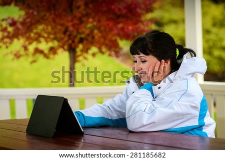 Women in the late twenties sitting with a tablet in the wooden arbor during the rainy autumn day