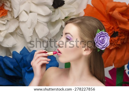 beautiful girl paints her lips with red lipstick and looks in the picture. Doing make up by herself with big summer flowers on bakcground