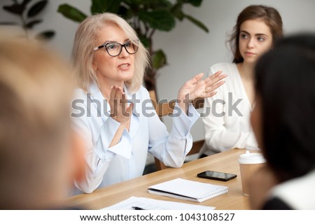 Photo of Attractive aged businesswoman, teacher or mentor coach speaking to young people, senior woman in glasses teaching audience at training seminar, female business leader speaker talking at meeting