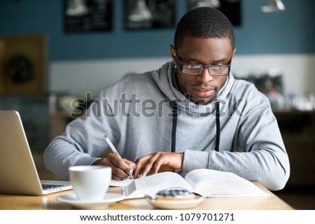 Focused millennial african american student in glasses making notes writing down information from book in cafe preparing for test or exam, young serious black man studying or working in coffee house Сток-фото © 