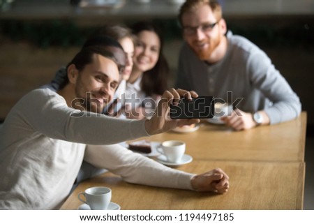 Photo of Millennial guy holding smartphone making self-portrait picture at friendly meeting with colleagues in café, happy friends smile for selfie sitting at coffee table, student shot photo with cellphone