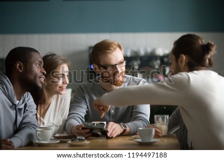 Photo of Young red haired guy show pictures on camera to friends, sitting in coffeeshop together having fun, millennial man share memories presenting photos on device to excited colleagues meeting in café