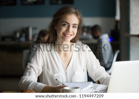 Photo of Portrait of smiling millennial student looking in camera studying in café, happy young female posing for picture working at computer, doing homework in coffeeshop, girl busy preparing report out