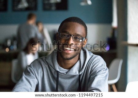 Photo of Portrait of smiling African American student looking at camera sitting in café, black millennial man posing making picture in coffeeshop, afro male in glasses drinking coffee working in coffeehouse