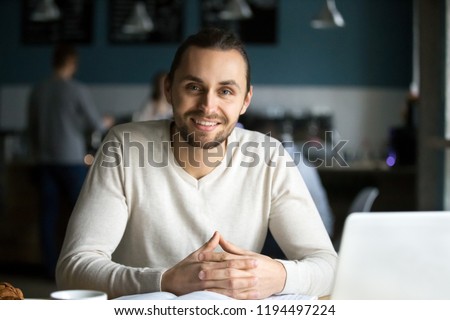 Photo of Portrait of smiling millennial man sitting in café with laptop and books on table, happy young guy work in coffeeshop using computer, male student look at camera busy preparing report in coffeehouse