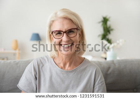 Photo of Smiling middle aged mature grey haired woman looking at camera, happy old lady in glasses posing at home indoor, positive single senior retired female sitting on sofa in living room headshot portrait