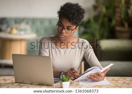 Focused young african american businesswoman or student looking at laptop holding book learning, serious black woman working or studying with computer doing research or preparing for exam online Сток-фото © 
