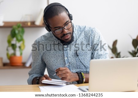 Focused african business man in headphones writing notes in notebook watching webinar video course, serious black male student looking at laptop listening lecture study online on computer e learning Foto d'archivio © 