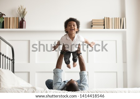 Happy funny african american kid boy flying in fathers arms looking at camera in bedroom, loving family single black dad holding lifting cute little child son playing plane bonding having fun on bed